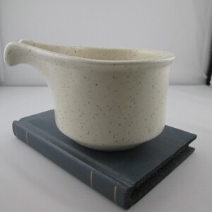 May include: A cream-colored ceramic gravy boat with a speckled finish and a single handle. The vessel features a wide, rounded bowl shape with a prominent pouring spout on one side, showcasing a simple, functional design for serving sauces.