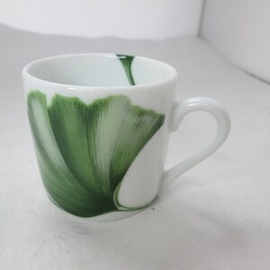 May include: White ceramic mug with a green ginkgo leaf design. The mug has a rounded shape and a handle.