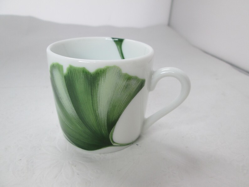 May include: White ceramic mug with a green ginkgo leaf design. The mug has a rounded shape and a handle.