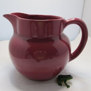 May include: A deep red ceramic pitcher with a rounded body and a flared lip. The pitcher has a handle and a smooth, glossy finish.