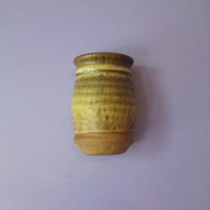 May include: A small, handcrafted ceramic vase with a cylindrical shape. The vase features a textured surface with horizontal bands of yellow, brown, and tan hues. The base and rim are a darker brown, creating a rustic, earthy aesthetic.