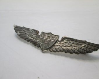 May include: A silver-toned pilot's wing pin with a shield in the center. The shield has the text "UNITED STATES ARMY AIR FORCES" on it.