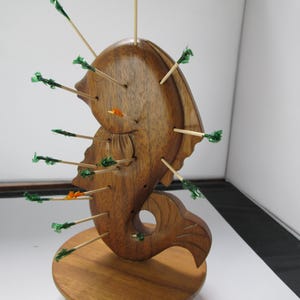 May include: A wooden seahorse-shaped toothpick holder. The holder is made of a light brown wood and has multiple holes with toothpicks inserted. The toothpicks have green and orange decorative tips. The holder sits on a round wooden base.