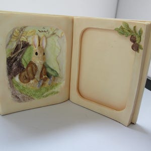 May include: Cream-colored, open book-shaped photo frame with a 3D relief of a rabbit in a forest scene on the left page. The right page has a recessed area for a photo. A sprig of leaves and berries adorns the top right corner.