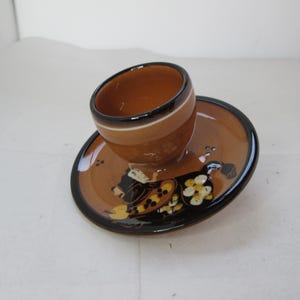May include: A brown ceramic egg cup with a black rim and a painted design of a woman in a black dress and yellow flowers. The egg cup is sitting on a matching brown ceramic saucer with a black rim.