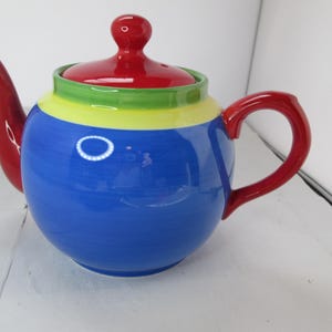 May include: A blue ceramic teapot with a red lid and handle. The teapot is decorated with stripes of green, yellow, and red.