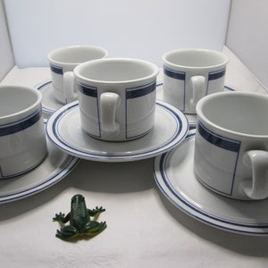 May include: Set of four white ceramic teacups with blue trim and matching saucers. The cups and saucers have a subtle textured design. A green plastic frog is in the foreground.