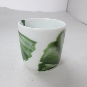 May include: A white ceramic cup with a green leaf design. The cup is approximately 7.5 cm tall and 6.5 cm in diameter.