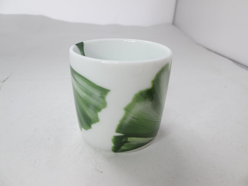 May include: A white ceramic cup with a green leaf design. The cup is approximately 7.5 cm tall and 6.5 cm in diameter.