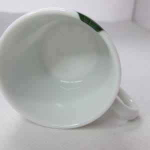 May include: White ceramic coffee cup with a green and white design. The cup has a handle and is empty.