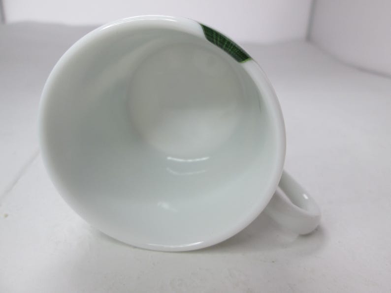 May include: White ceramic coffee cup with a green and white design. The cup has a handle and is empty.