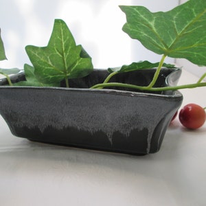 May include: A black ceramic rectangular dish with a white glaze. The dish is sitting on a white surface with green ivy leaves and red cherries.