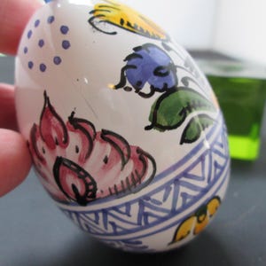 May include: A hand-painted ceramic egg with floral designs in pink, blue, yellow, and green. The egg has a white base with a blue and white geometric pattern around the bottom. A green glass cube is in the background.