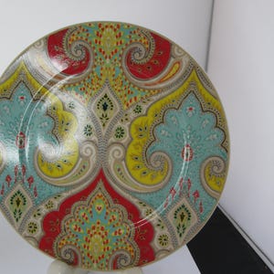 May include: A round decorative plate with a colorful paisley design. The plate features a repeating pattern of teal, yellow, red, and gray swirls and floral motifs. The plate is likely made of ceramic or porcelain and is suitable for serving food or as a decorative item.