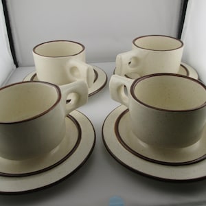 May include: A set of four off-white ceramic cups and matching saucers, each featuring a speckled finish and a dark brown rim. The cups have a thick, rounded handle design, and the saucers have a circular indentation to hold the cup securely.