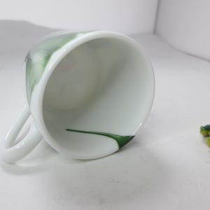 May include: White ceramic teacup with a green floral design. The inside of the cup is white with a green line.