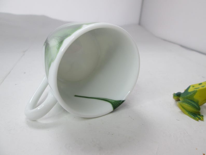 May include: White ceramic teacup with a green floral design. The inside of the cup is white with a green line.