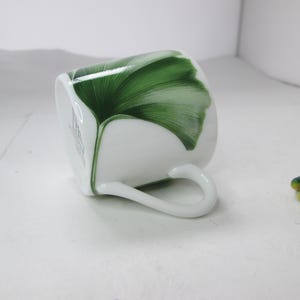 May include: White porcelain teacup with a green ginkgo leaf design. The cup has a handle and is tilted on its side.