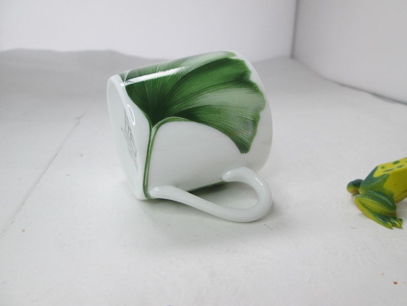 May include: White porcelain teacup with a green ginkgo leaf design. The cup has a handle and is tilted on its side.