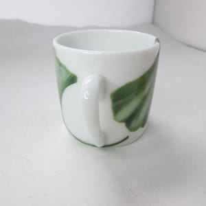 May include: White ceramic mug with a green and white ginkgo leaf design. The mug has a rounded handle.