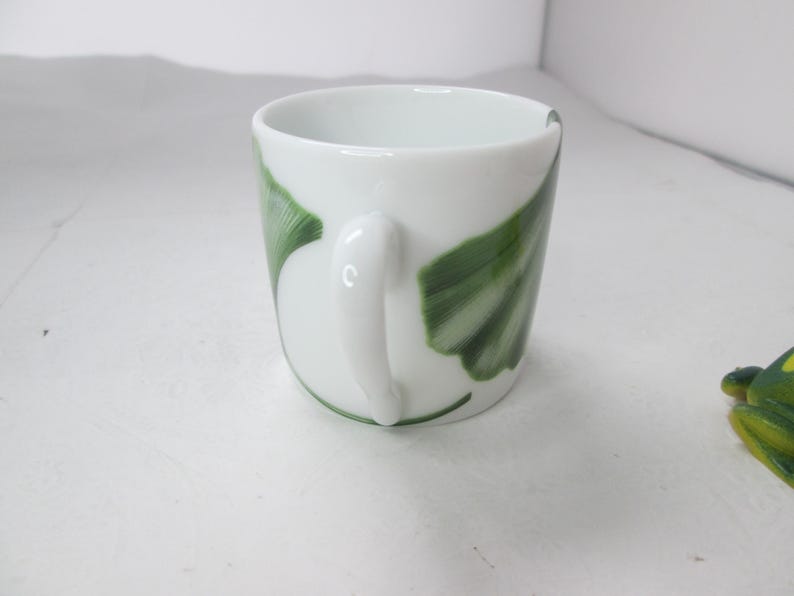 May include: White ceramic mug with a green and white ginkgo leaf design. The mug has a rounded handle.