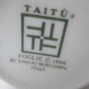 May include: A white ceramic plate with a green logo that reads "Tait&ugrave;" with a stylized symbol and the text "Foglie &copy; 1996 by Emilio Bergam&iacute;n Italy".