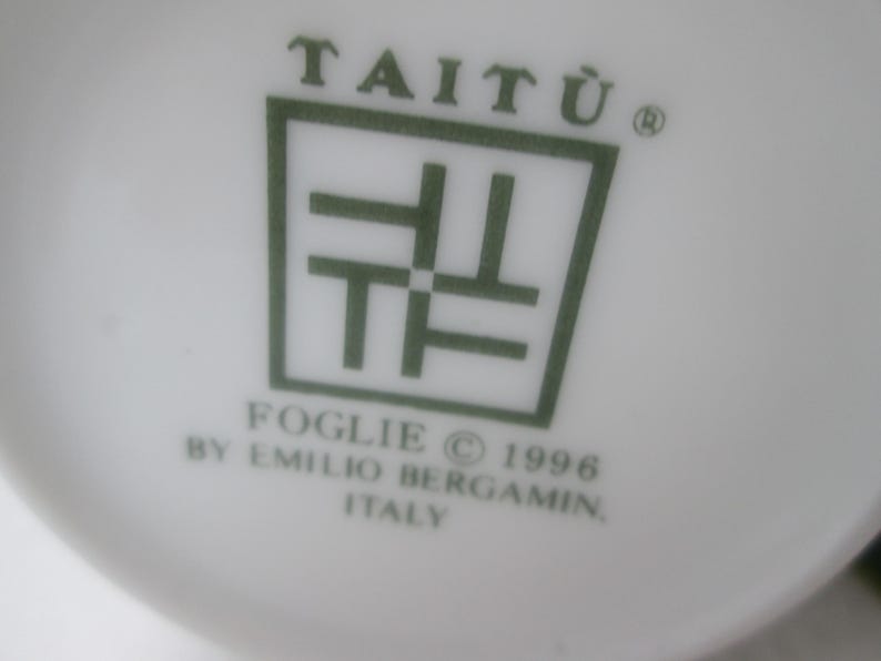 May include: A white ceramic plate with a green logo that reads "Tait&ugrave;" with a stylized symbol and the text "Foglie &copy; 1996 by Emilio Bergam&iacute;n Italy".