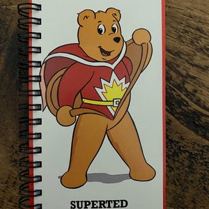 May include: A red and yellow superhero bear with a cape and a belt, standing with a smile. The text "SUPERTERD" is printed below the bear.