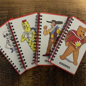 May include: Four small spiral bound notebooks with colorful cartoon characters on the covers. The covers feature a skeleton, a spotted figure, a cowboy, and a superhero bear. The notebooks are labeled "Space", "Team", "Superted", and "Superted".