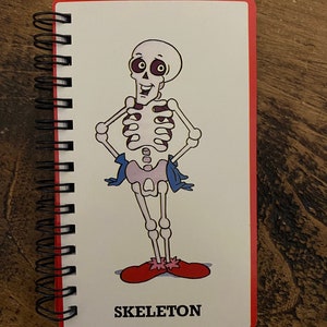 May include: A spiral bound notebook with a red cover and a cartoon skeleton illustration. The skeleton is wearing red shoes and a blue sash. The text "SKELETON" is printed below the illustration.