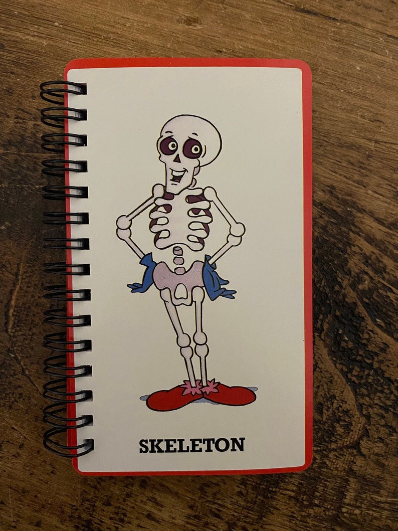May include: A spiral bound notebook with a red cover and a cartoon skeleton illustration. The skeleton is wearing red shoes and a blue sash. The text "SKELETON" is printed below the illustration.