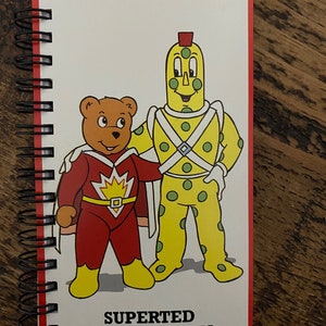 May include: A red and yellow notebook with a spiral binding. The cover features a cartoon illustration of a red superhero bear with a yellow cape and a yellow and green spotted character with a red hat. The text "Superted & Spottyman" is printed in black at the bottom of the cover.