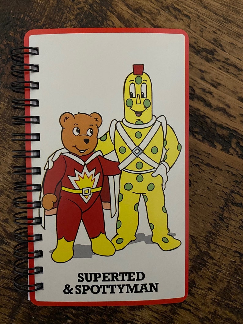May include: A red and yellow notebook with a spiral binding. The cover features a cartoon illustration of a red superhero bear with a yellow cape and a yellow and green spotted character with a red hat. The text "Superted & Spottyman" is printed in black at the bottom of the cover.