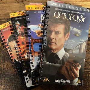 May include: Three spiral-bound notebooks featuring James Bond film covers. The front notebook displays the Octopussy movie poster with Roger Moore holding a gun, the title, and the text 007 Wide Screen Collection. The other two notebooks show partial covers.