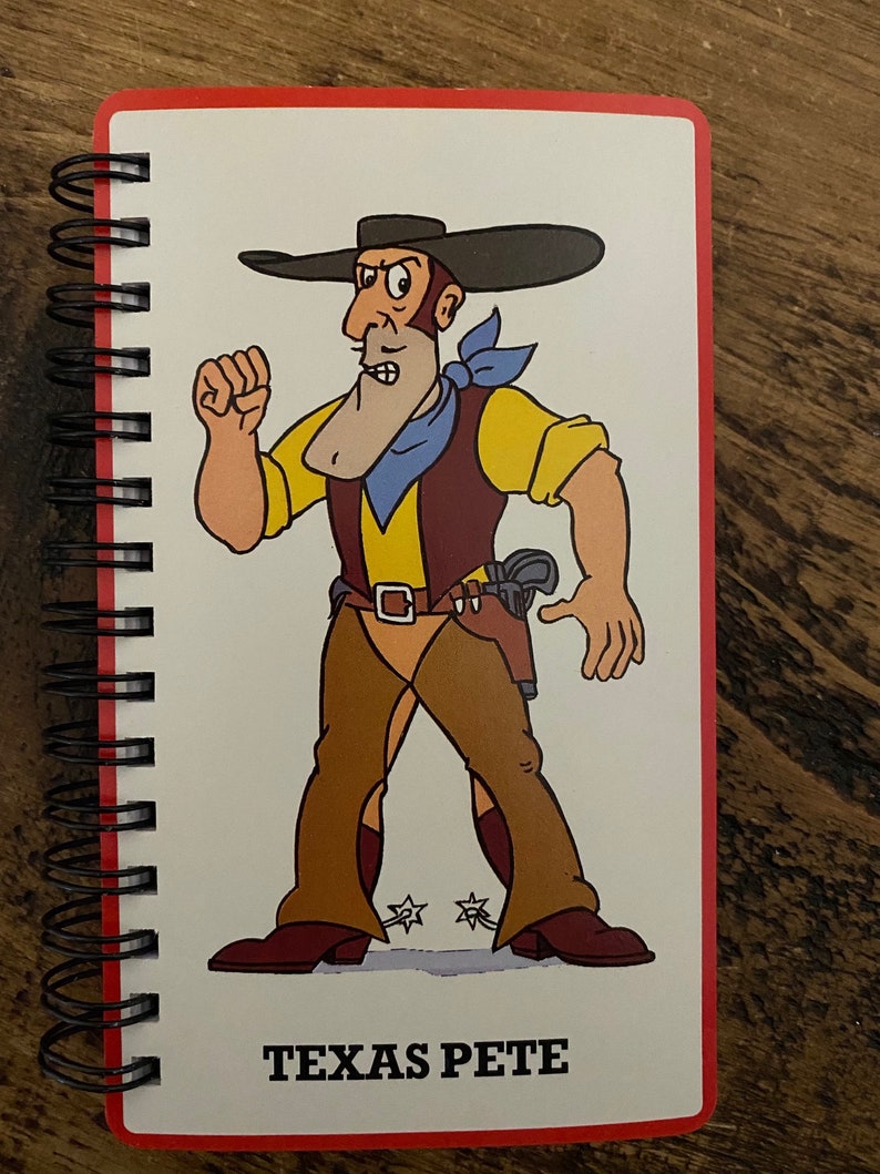 May include: A red spiral bound notebook with a cartoon illustration of a cowboy wearing a brown hat, yellow shirt, blue bandana, and brown pants with spurs. The cowboy is holding a gun and has a stern expression. The text "TEXAS PETE" is printed below the illustration.