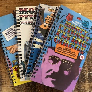 May include: A stack of four spiral bound notebooks with colourful covers featuring images and text related to Monty Python's Flying Circus. The top notebook has a blue cover with the text "Monty Python's Flying Circus" in white and red letters. The text "Series 2 Episodes 11-13" is in white letters on a blue background. The text "Featuring SPAM, SPAM, SPAM, SPAM, the Dirty Hungarian Phrasebook and far too much cannibalism" is in white letters on a blue background. The number "15" is in white letters on a red circle.