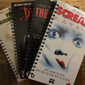 May include: A set of four spiral-bound notebooks featuring horror movie poster covers. The top notebook displays the Scream film poster with a blue-toned face and red text. Other visible titles include The Frighteners and Dracula, all bound with black plastic coils.