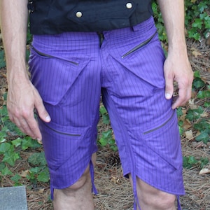 May include: A person wearing a black sleeveless vest with gold buttons and a black hat with a studded design. They are also wearing purple striped shorts.