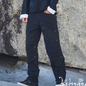 May include: A person wearing a black button-down shirt with silver studs and white cuffs, paired with black pants. The outfit includes a patterned cap and black sneakers. The clothing has a unique design.