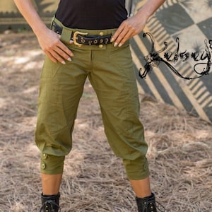May include: A woman wearing a black, long-sleeved shirt with lace detailing, olive green pants, a brown hat, and black boots. The pants have a belt with gold accents. The woman is standing in front of a striped background.
