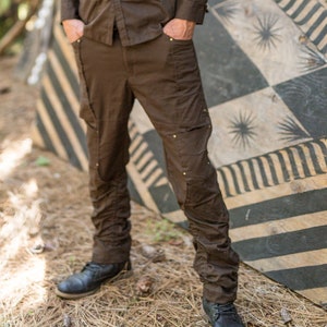 May include: A man wearing a brown shirt and brown pants stands in front of a black and white striped wall. He is wearing a black hat with a design on it.