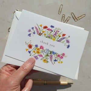 May include: White thank you cards featuring a floral design in a variety of colours, including red, yellow, purple, and blue. The words "thank you" are printed in a white rectangle. Gold paper clips and a pen are in the background.