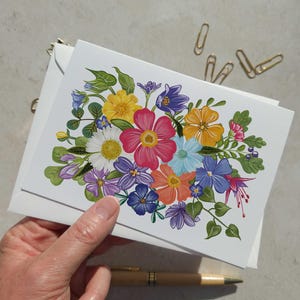 May include: A greeting card with a vibrant floral design, showcasing pink, yellow, blue, and orange flowers. The card is held with a white envelope. Gold paper clips and a pen are also visible.