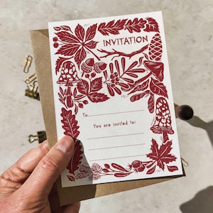 May include: A rectangular white invitation card featuring a dark red botanical print of leaves, acorns, mushrooms, and pinecones. The text reads "INVITATION", "To:", and "You are invited to:" with dotted lines for writing details, tucked into a brown envelope.