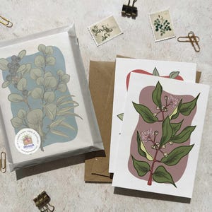 May include: A set of blank cards and envelopes featuring botanical illustrations. One card displays eucalyptus leaves on a blue background, while others showcase floral designs on a white backdrop. The cards are packaged with brown envelopes and decorative paper.
