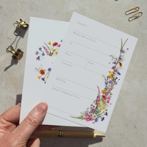 May include: White invitation cards with watercolour floral designs. One card features a floral wreath, the other a floral border. The invitation includes fields for names, date, time, location, and RSVP information.