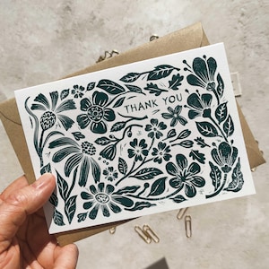 May include: A white rectangular greeting card featuring a dark green block-print style floral pattern and the text THANK YOU printed in the center. The card is positioned against a brown paper envelope, showcasing a detailed botanical illustration.