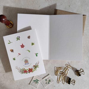 May include: A greeting card with a watercolor floral design, featuring red poinsettias, berries, and greenery. The card is open, revealing a blank interior. Additional stationery items include paper clips, stamps, and a wooden stamp.