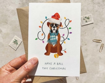 May include: A Christmas card featuring a Boxer dog wearing a Santa hat and a blue scarf, surrounded by colorful Christmas lights. The card reads "Have a ball this Christmas."