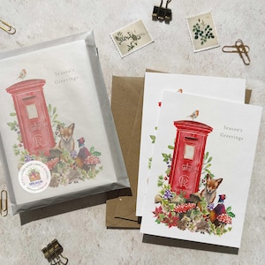 May include: Greeting cards featuring a red postbox design with a fox, pheasant, and other woodland creatures. The cards are packaged in a clear sleeve and include the text "Season's Greetings". Paperclips and stamps are scattered around the cards.
